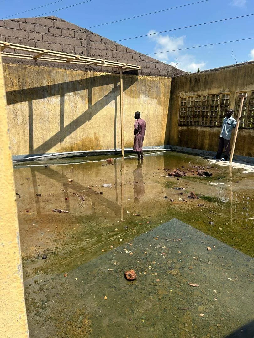 Two people stand in a flooded, unfinished concrete courtyard beneath a bright blue sky.