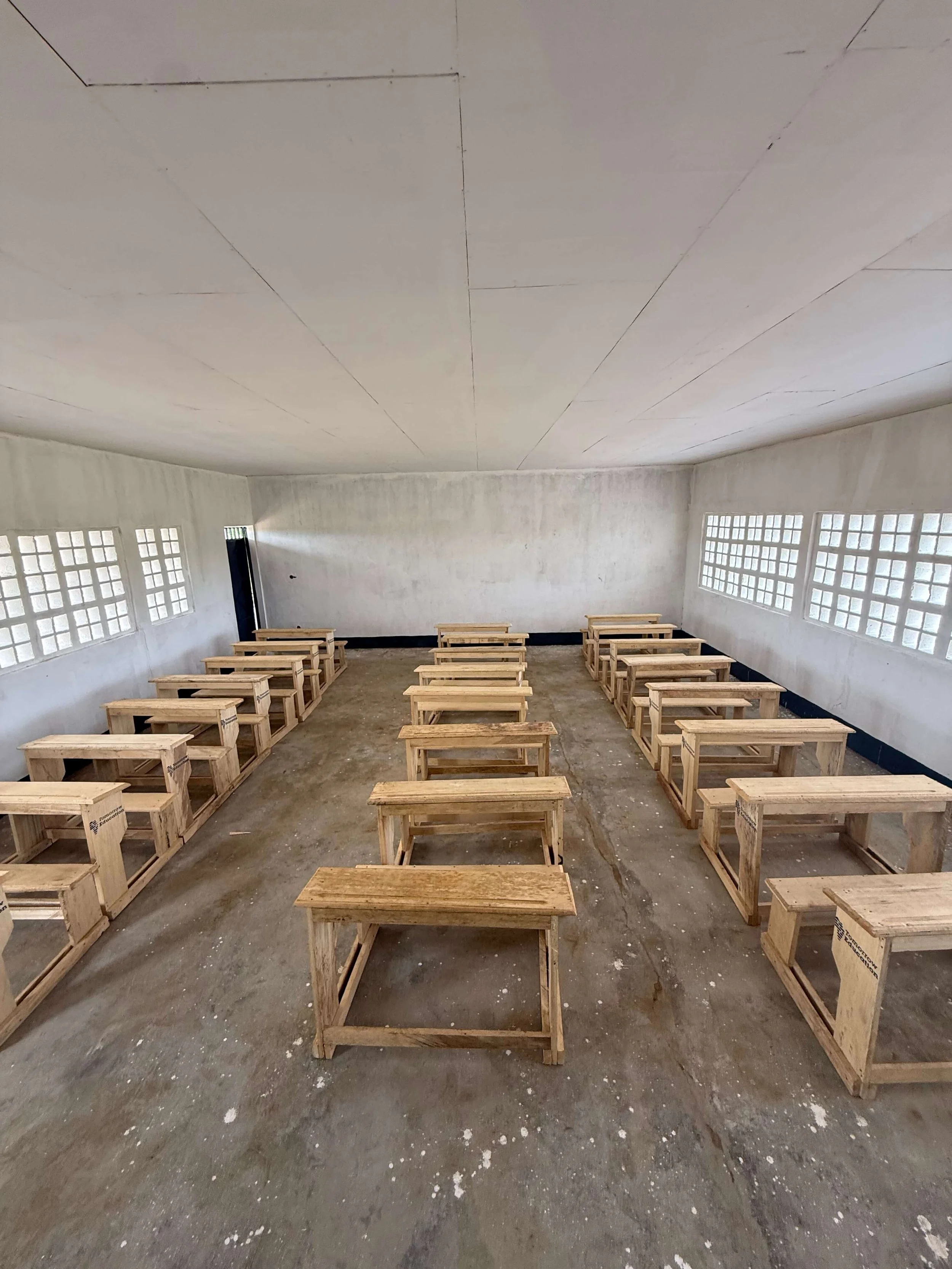 Empty classroom with rows of wooden desks, concrete walls, and glass-block windows.