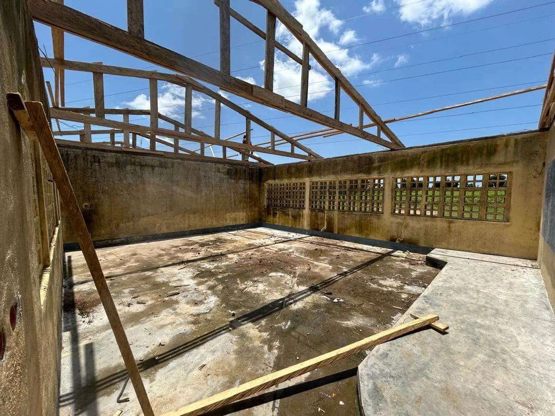 Concrete school building interior with exposed wooden roof framing, ventilation-block windows, and debris-covered floor during construction