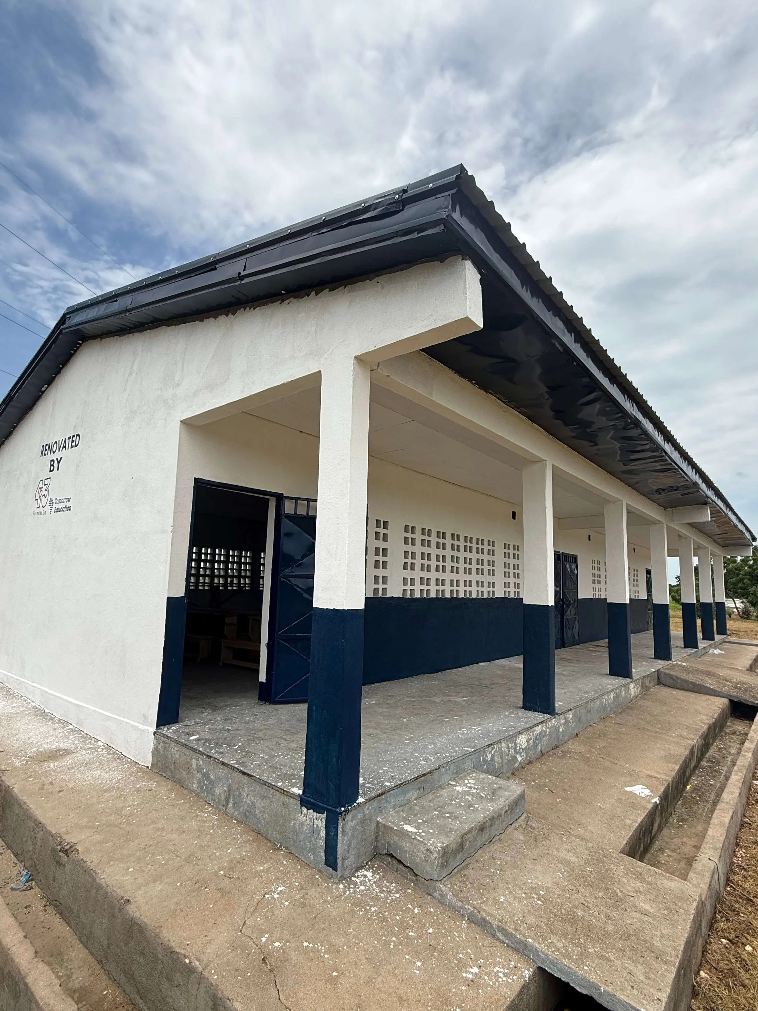 Renovated school building with white walls, navy-blue columns, covered walkway, and concrete drainage channel under a cloudy sky