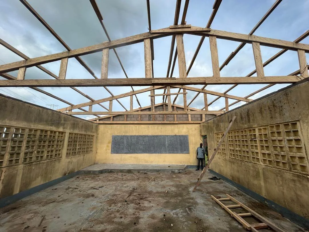 Interior of a partially demolished building with exposed wooden roof framing, concrete block windows, and a person standing near a chalkboard.