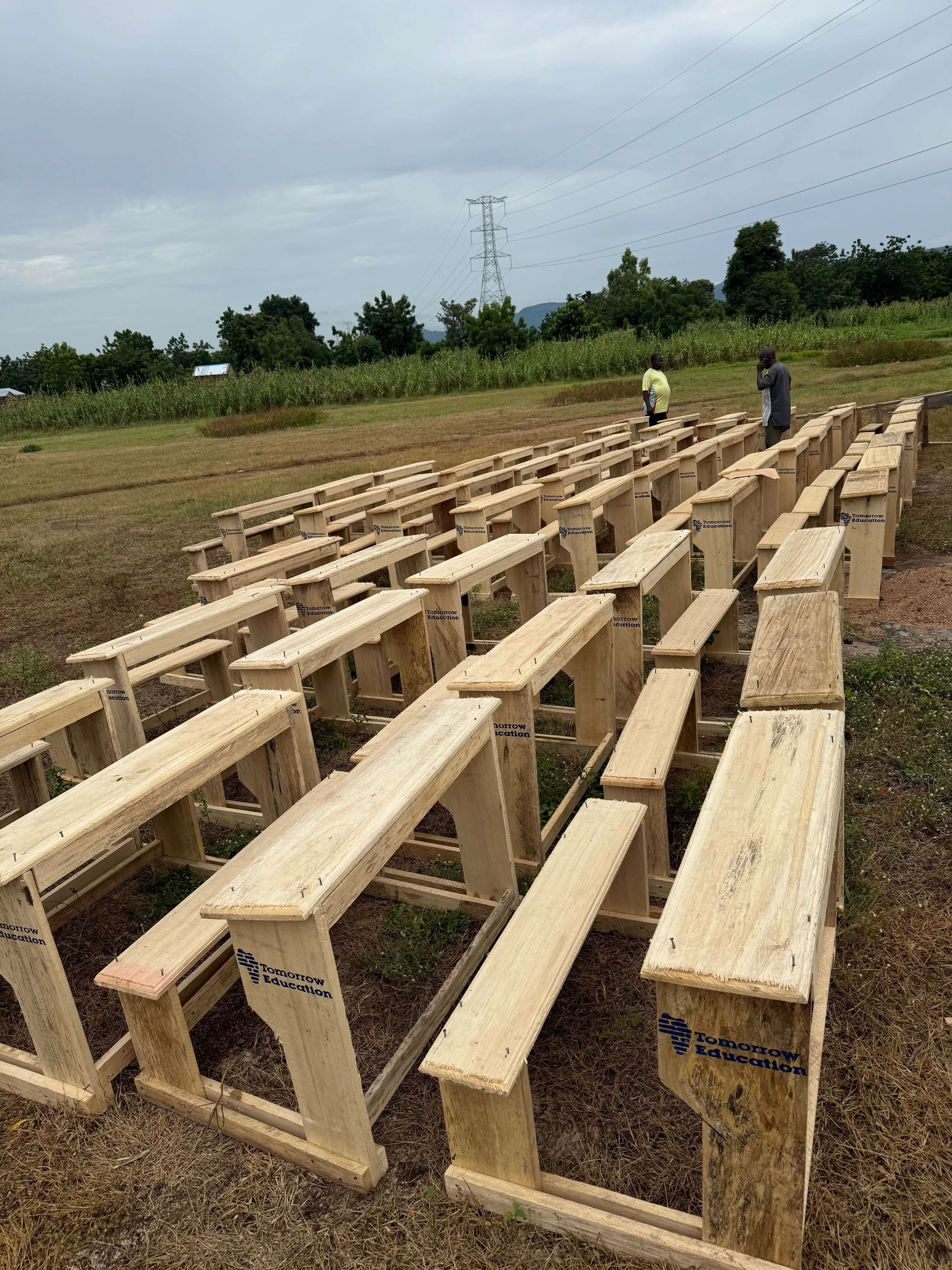 Rows of handmade wooden school desks labeled “Tomorrow Education” in a grassy field, with two people, crops, trees, mountains, and high-voltage power lines behind them.