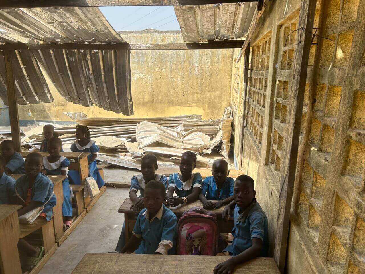 Schoolchildren in blue uniforms sit at wooden desks inside a damaged classroom with a partially collapsed corrugated metal roof.