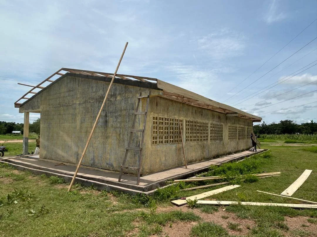 Partially constructed concrete building with exposed roof framing, ladder, wooden planks, and grassy surroundings