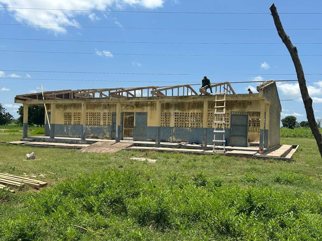 Workers repair the roof of a weathered concrete building beside a grassy field under a blue sky.