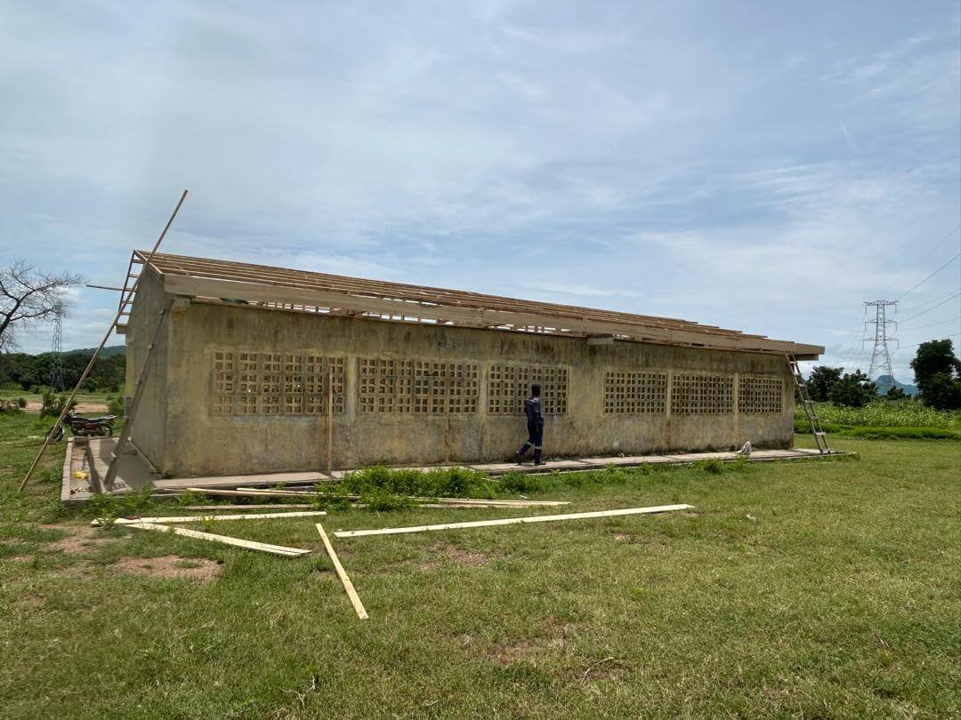 Unfinished concrete building with breeze-block windows, exposed roof framing, scattered lumber, and a person walking outside