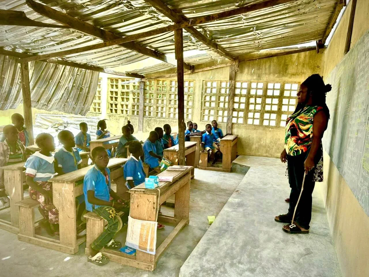Teacher leading a class of schoolchildren in a new classroom with wooden desks and a corrugated metal roof