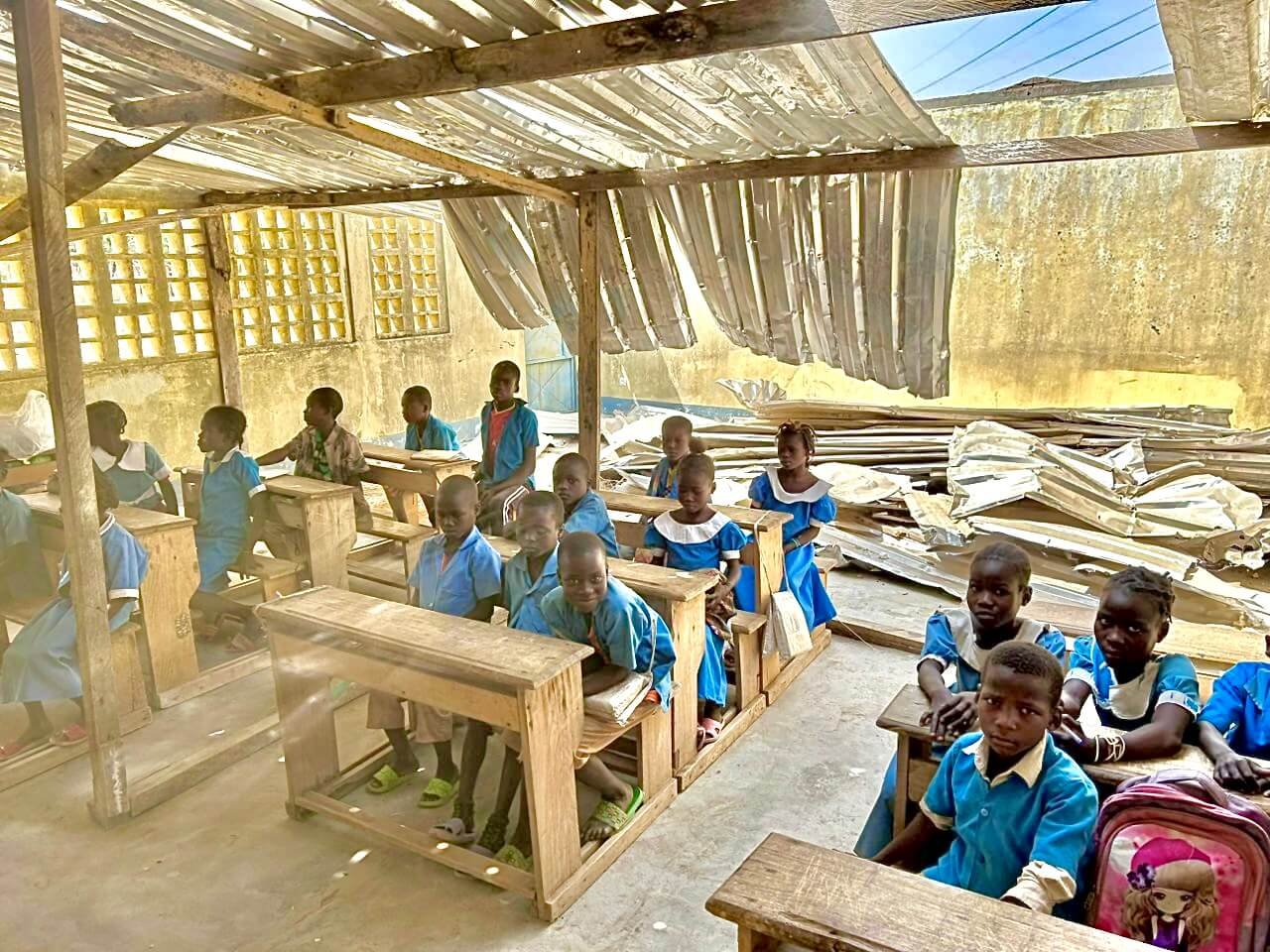 Schoolchildren in blue uniforms sit at wooden desks inside a classroom with a partially exposed corrugated metal roof.