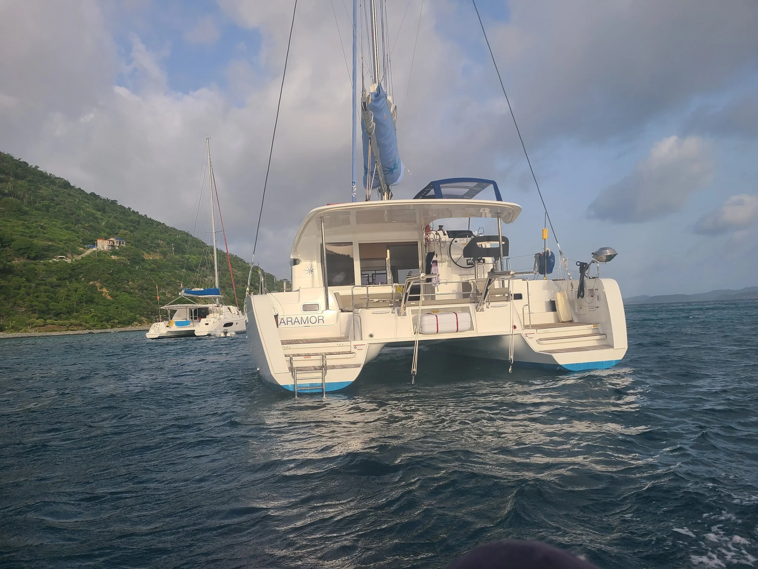 A white sailing yacht named 'Aramor' floating on the water near a shoreline with green hills and another boat visible in the background.