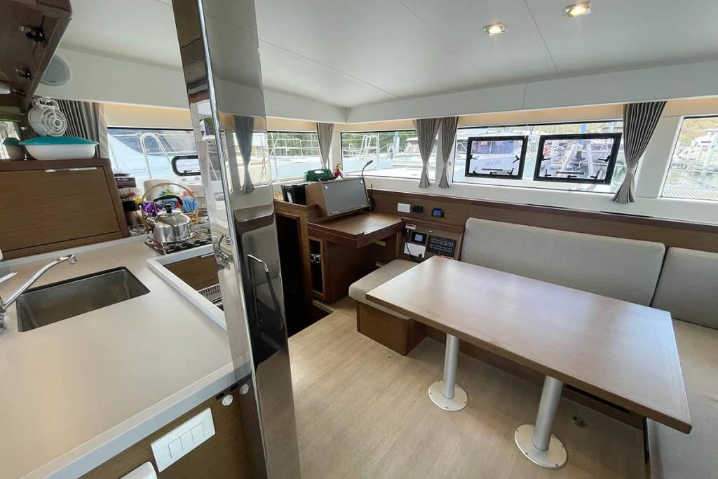 Interior view of a boat's living and kitchen area featuring a dining table with a cushioned bench, windows with curtains, a small kitchen with a sink and stovetop, and various controls and electronic equipment on the wall.