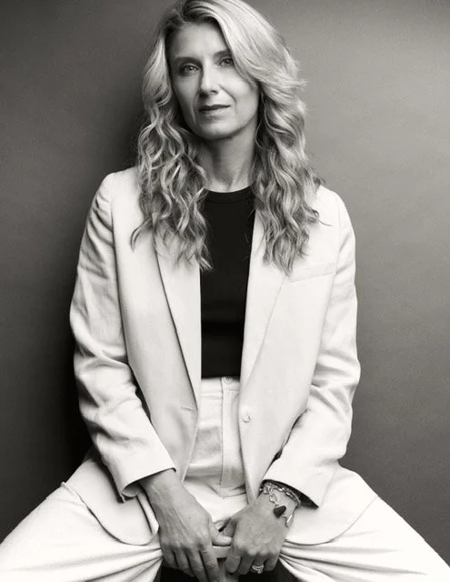 A woman with long wavy hair wearing a light-colored blazer and black top, sitting against a plain wall, looking at the camera.