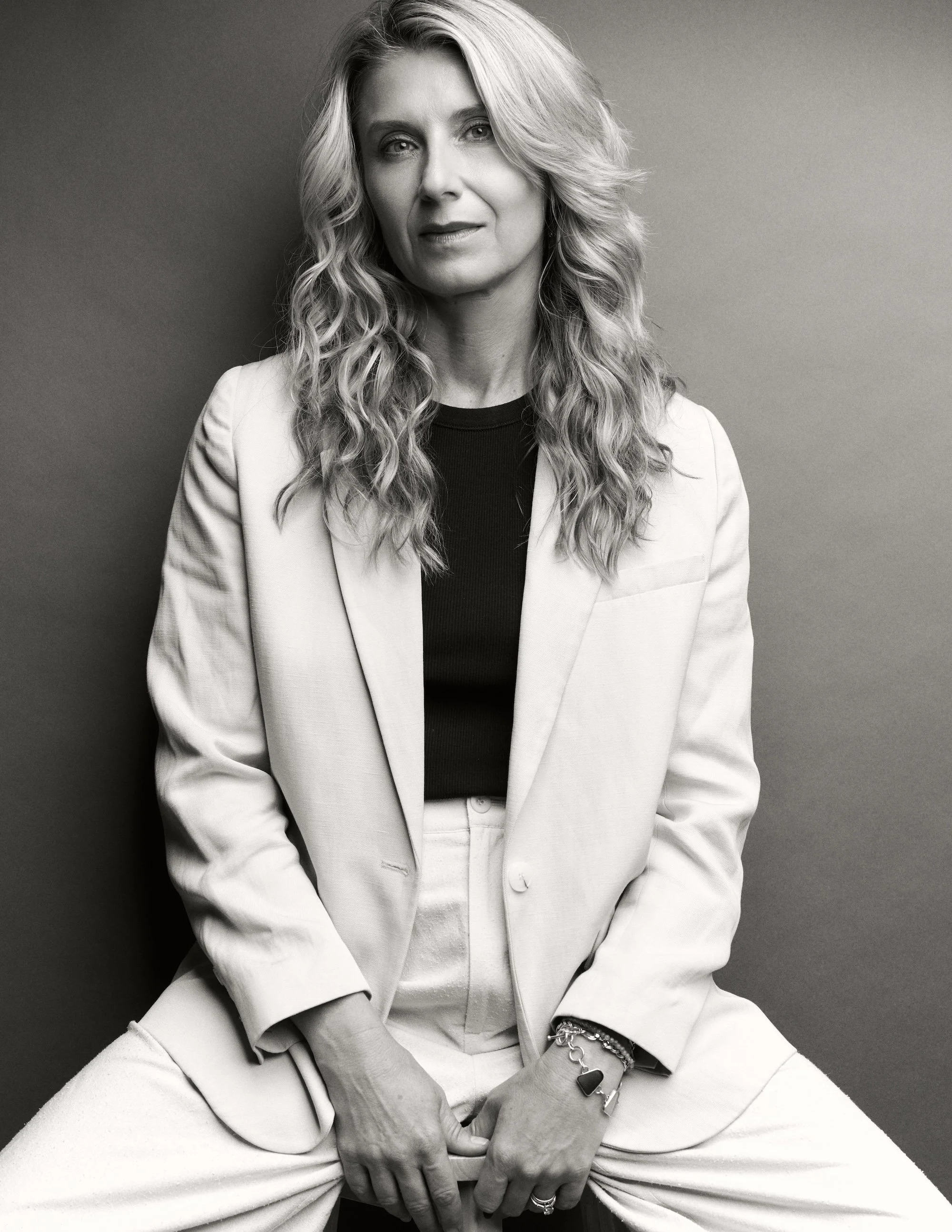Black and white portrait of a woman with wavy hair wearing a blazer and sitting with her hands resting on her knees.