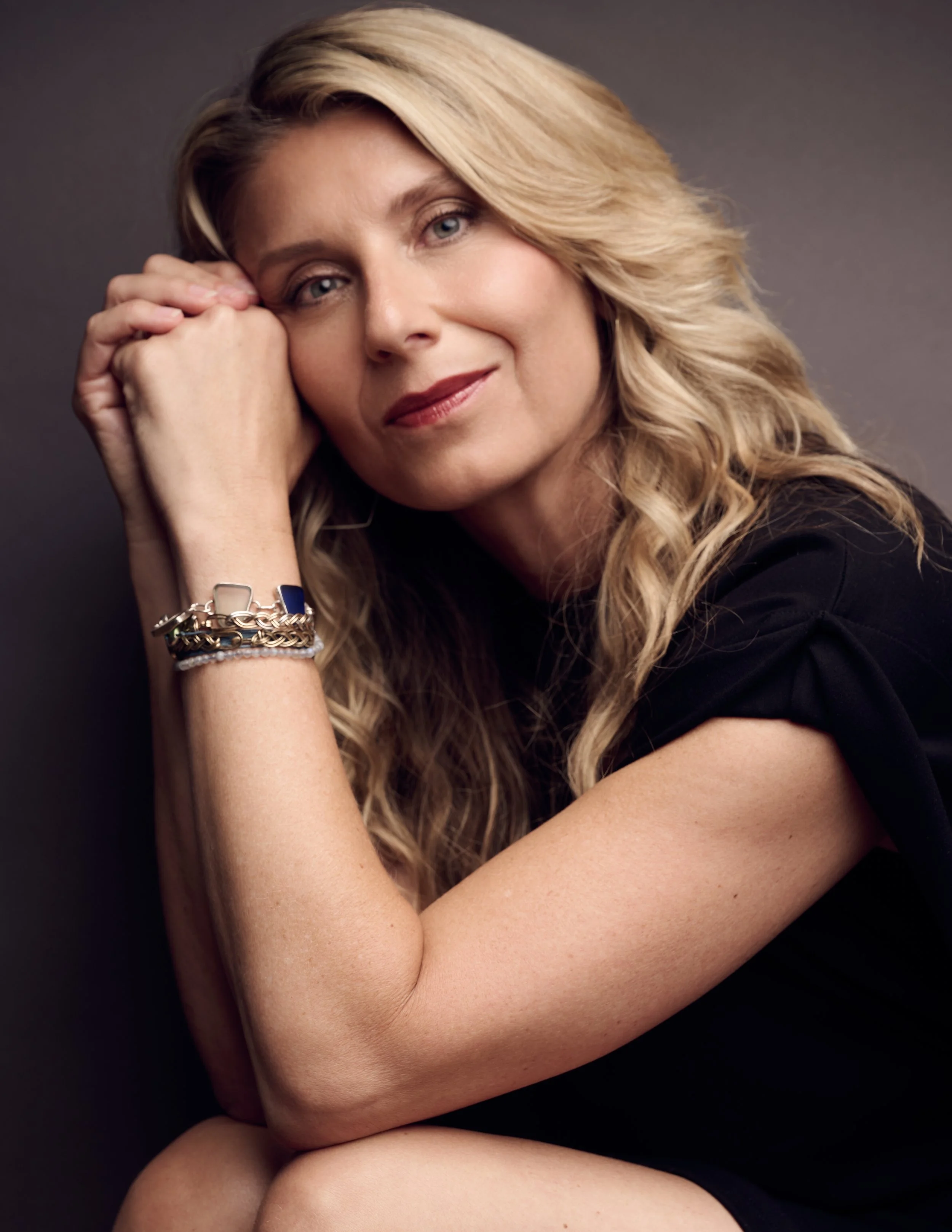 Close-up portrait of a woman with blonde wavy hair, smiling softly and resting her head on her hand against a dark background.