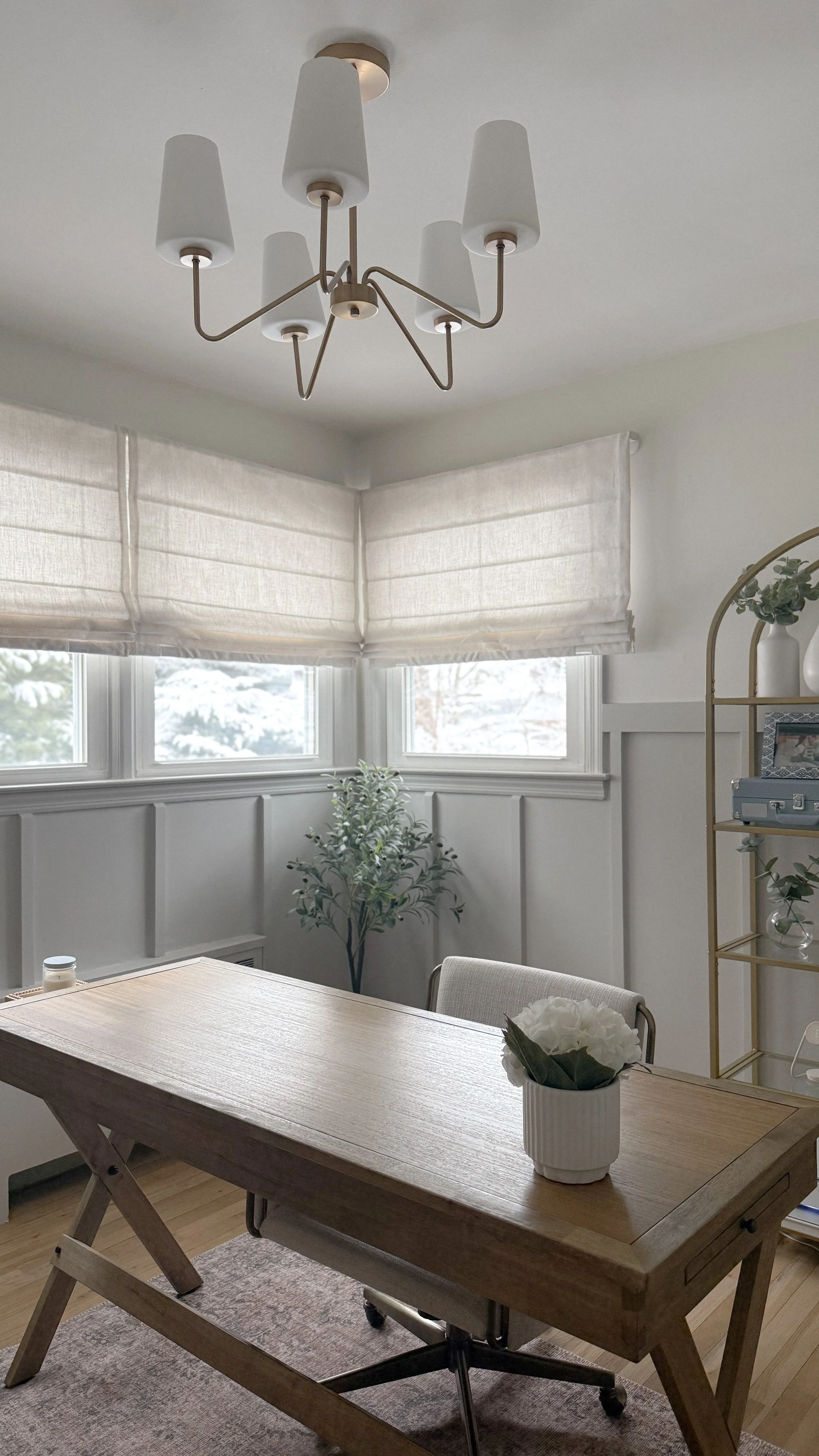 A dining room with a wooden table, white flower vase, a ceiling chandelier, windows with white curtains, and a shelf with vases and picture frames.