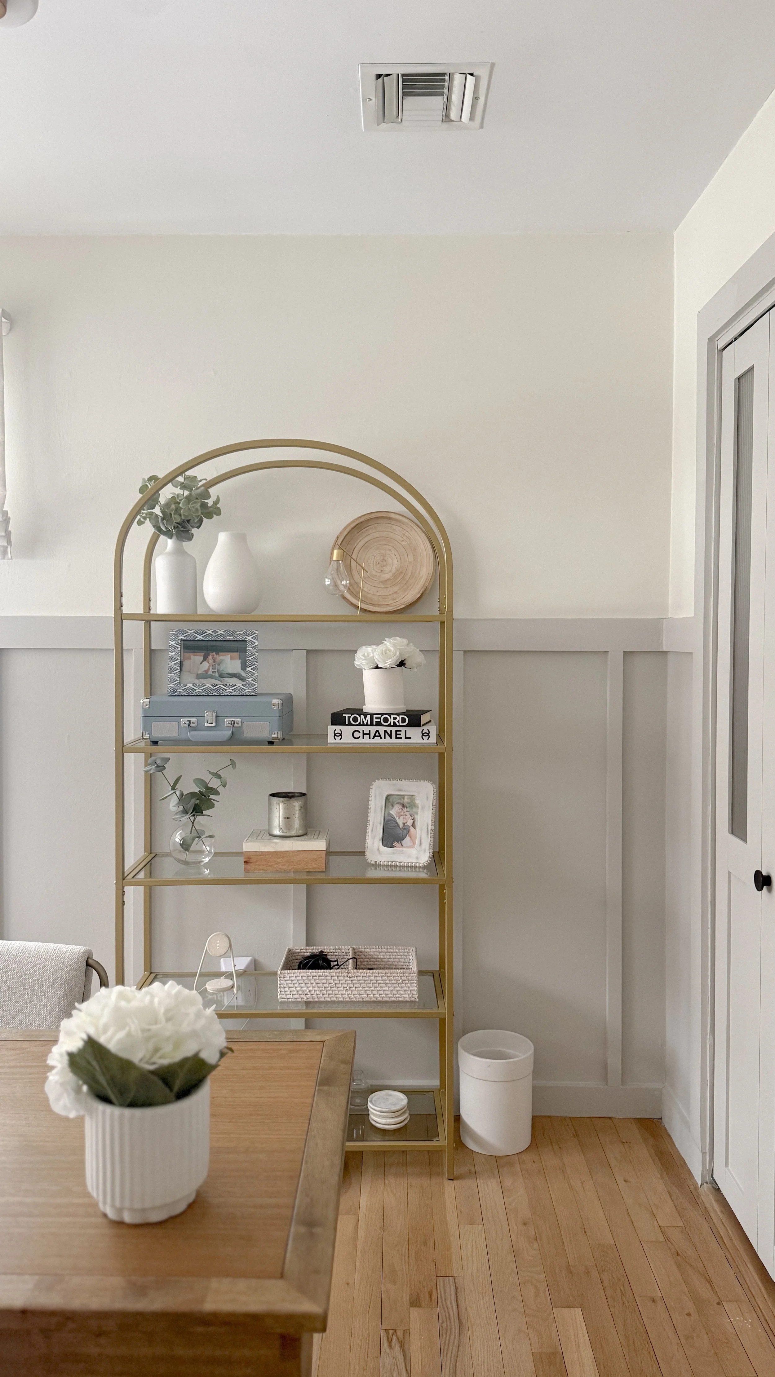 A gold-colored arched shelving unit with decorative items, picture frames, books, and small storage containers, situated against a white wall in a room with wooden flooring.
