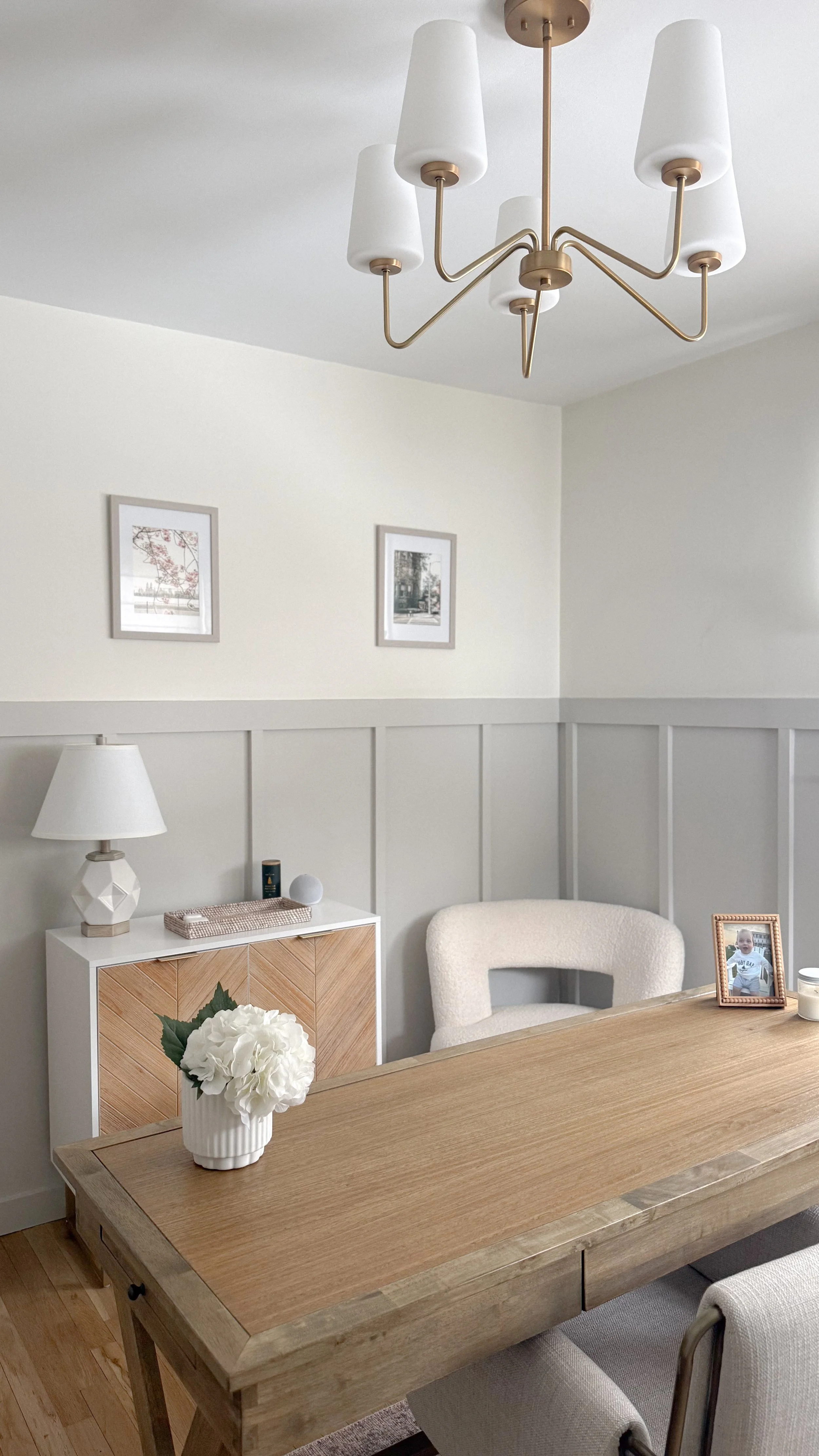A modern dining room with a wooden table, white chairs, a sideboard with a table lamp and decorative items, framed black-and-white photographs on the wall, a floral vase on the table, and a gold chandelier with white lampshades hanging from the ceili