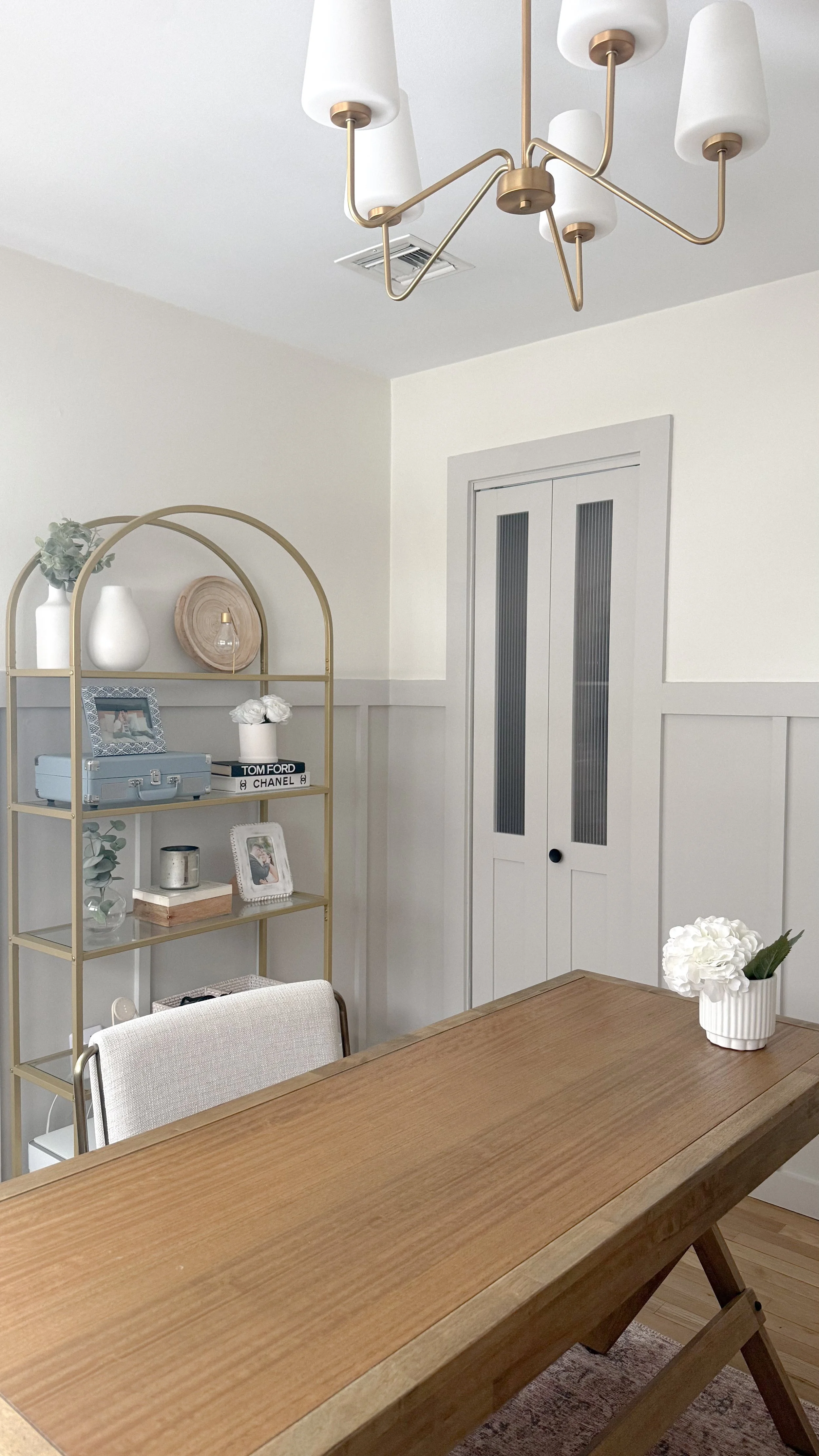 A dining room with a wooden table, a white flower vase, a gold shelving unit with decor, and a white door with glass panels.