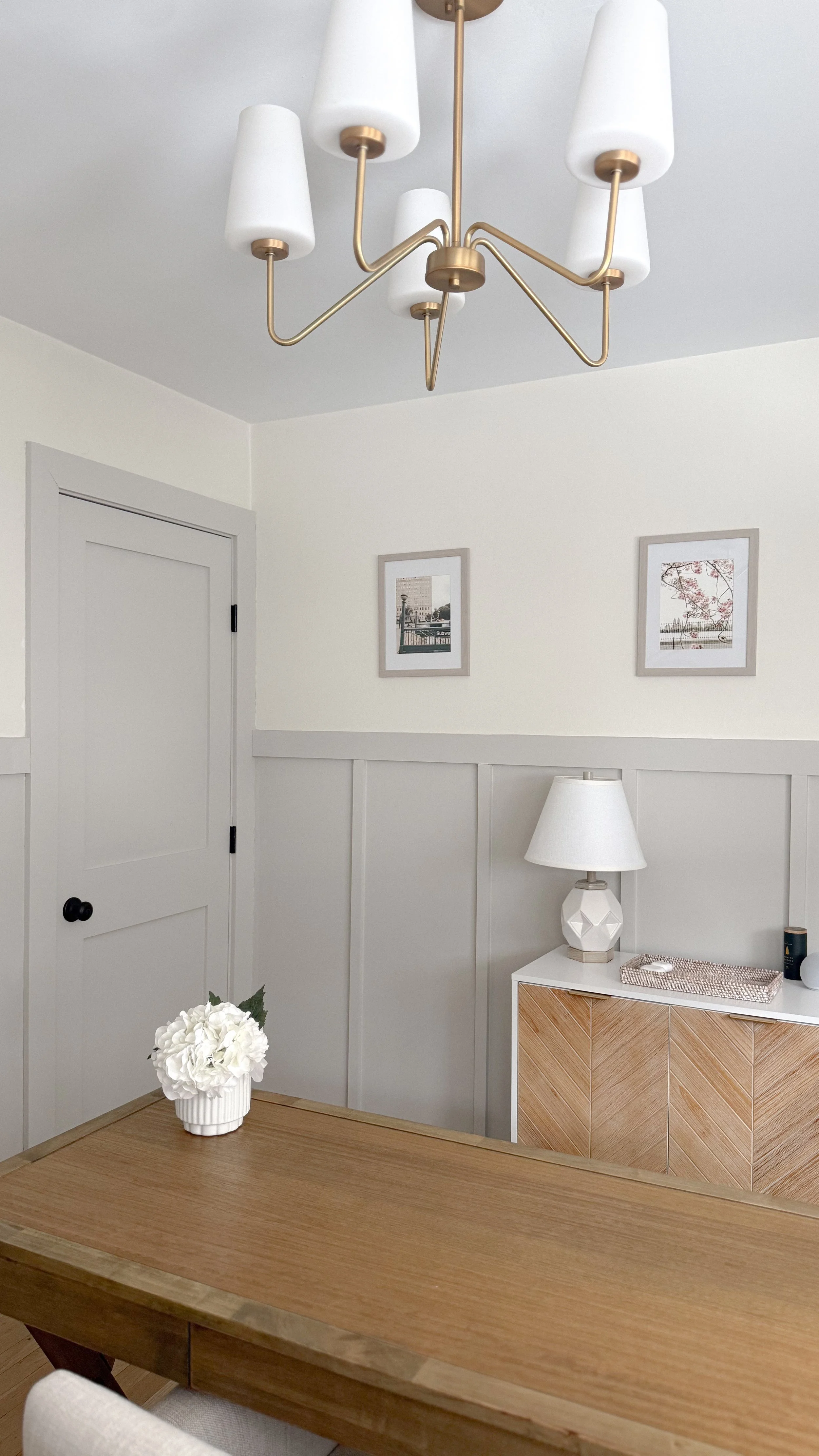 A room with a modern chandelier, framed pictures on the wall, a table with a white flower vase, and a sideboard with a white lamp.