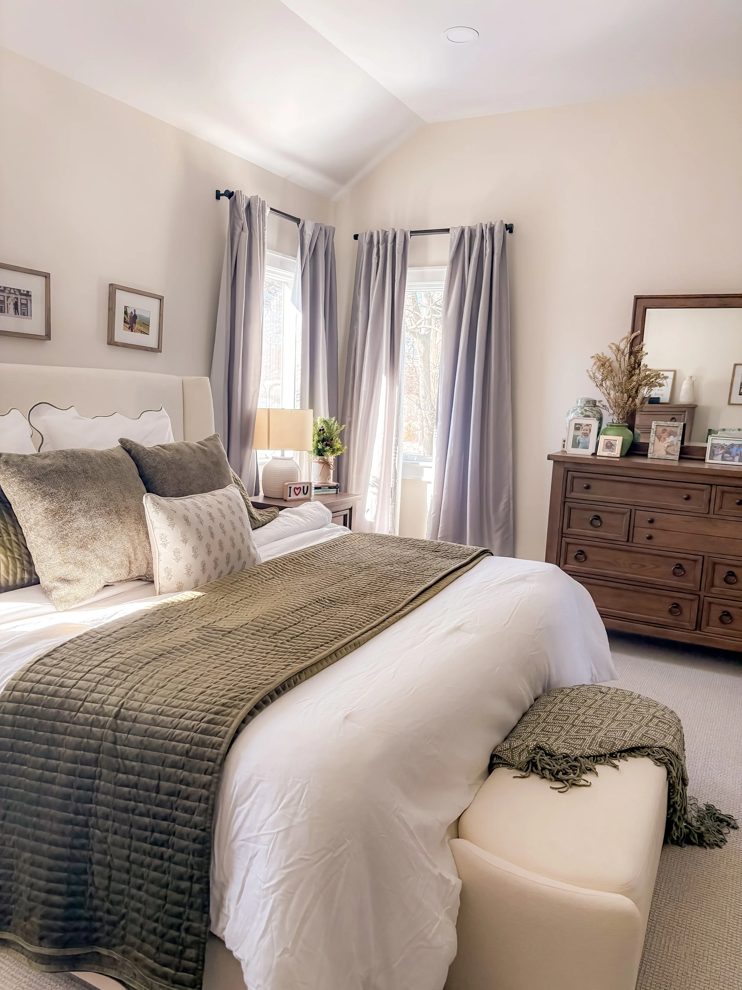 A cozy bedroom with a large bed covered in white linens, several pillows, and a quilted blanket. There is a small ottoman at the end of the bed with a green and beige throw. Two windows with light gray curtains allow natural light into the room. A wo