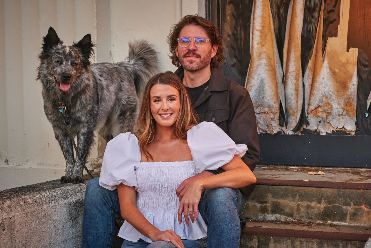 A smiling woman sitting on steps next to a man with glasses and long hair, with a speckled dog standing beside them. They are outdoors in front of a building with a textured surface and artwork.