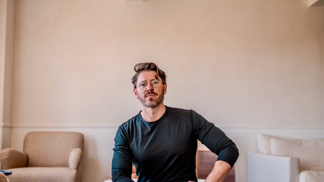 A man with glasses and a beard sitting in a beige armchair in a softly lit room with cream-colored walls.