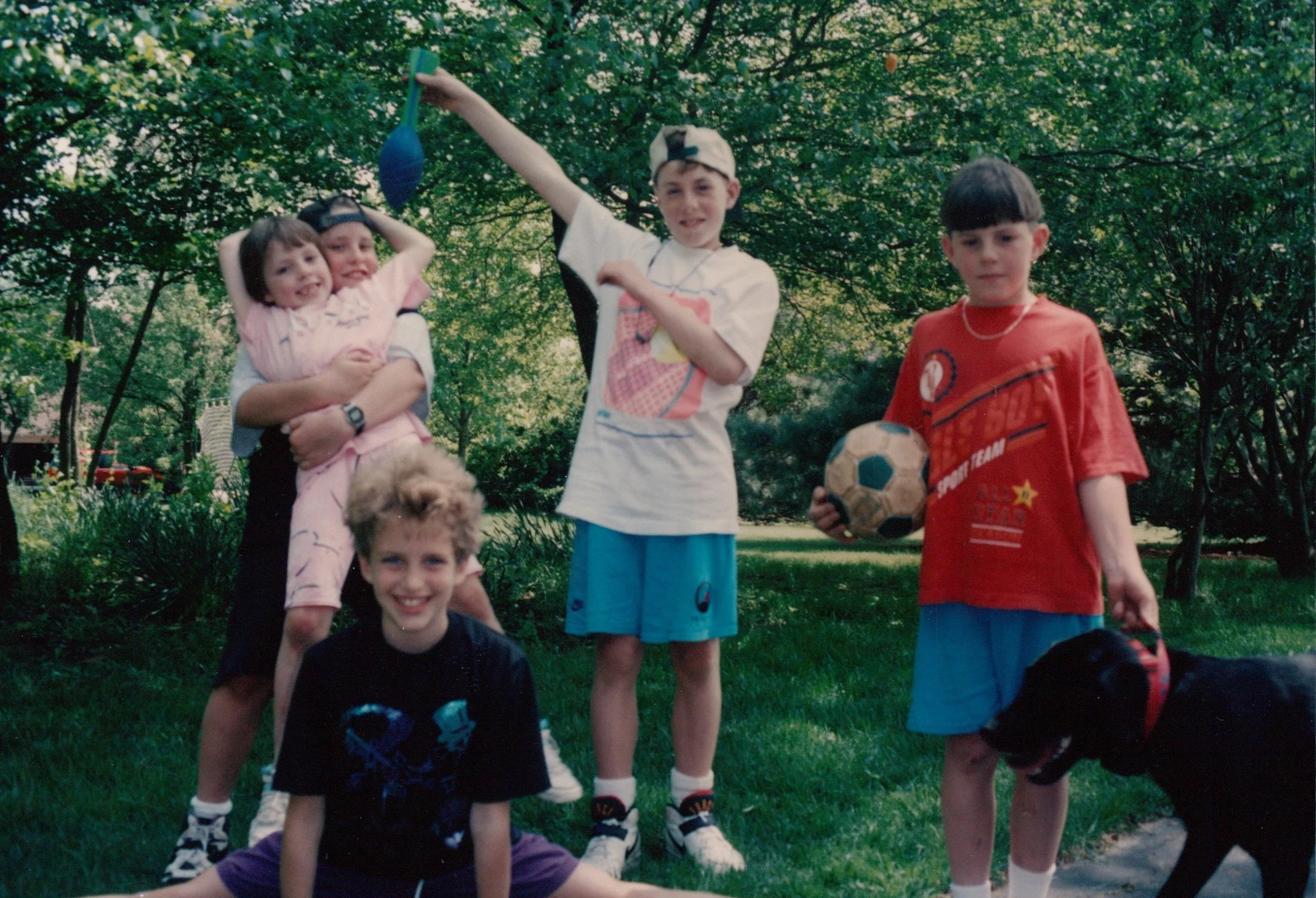 Group of five children and a black dog in a park, some holding soccer balls, surrounded by green trees.