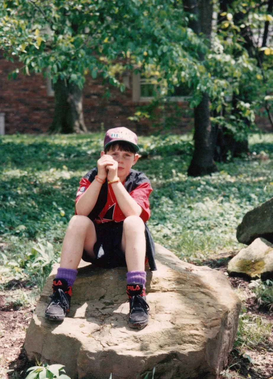 A young boy sitting on a large rock outdoors on a grassy area surrounded by trees and greenery, wearing a cap, red and black sports jersey, shorts, sneakers, and purple socks.