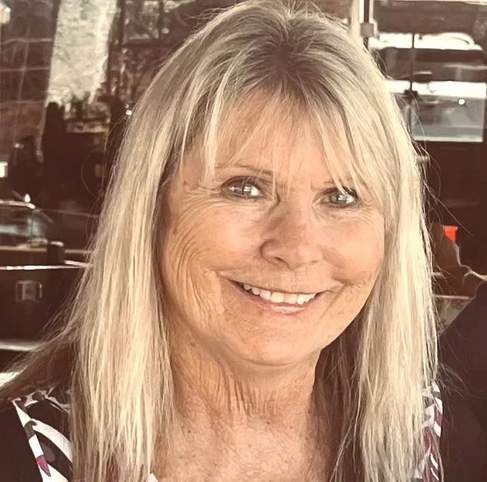 A smiling woman with long blonde hair and blue eyes, wearing a patterned top, indoors with a brick wall and cars visible in the background.