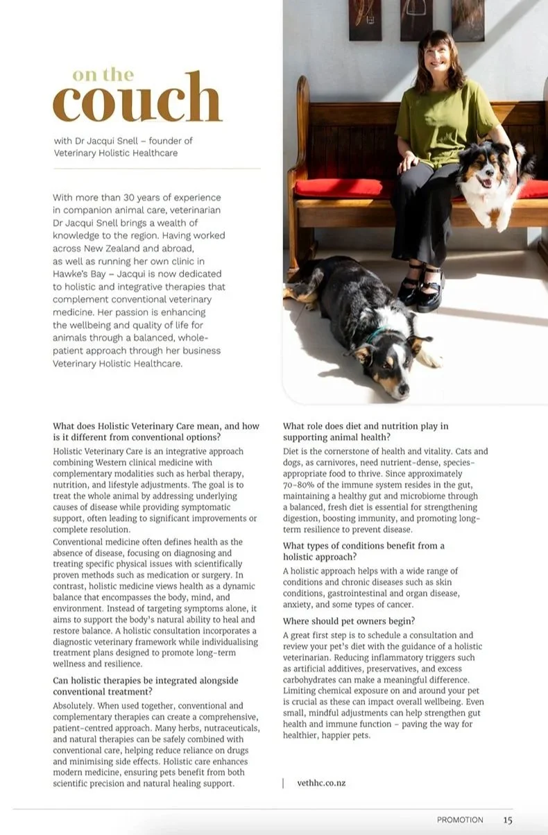 A woman sitting on a wooden bench with red cushions, smiling, with two dogs, one on her lap and one lying on the floor, in a room with gray walls and framed artwork.