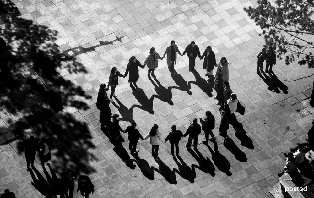 A group of people holding hands in a circle, casting long shadows on the ground in an outdoor setting with trees nearby.