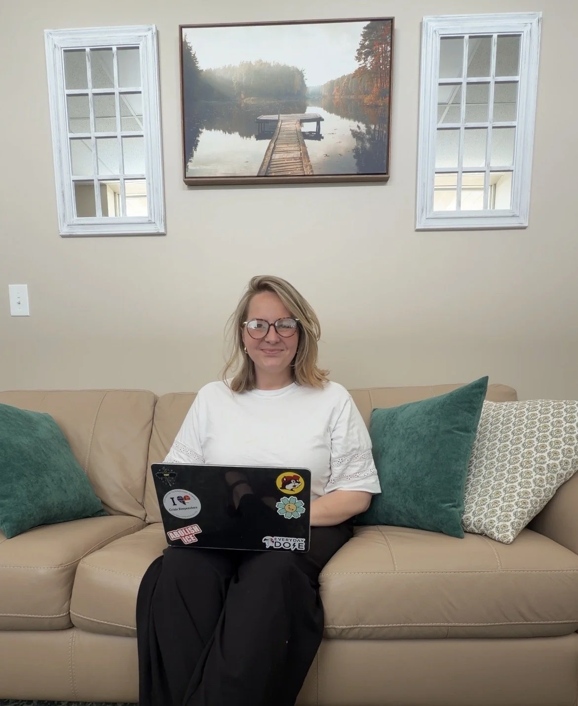 A woman with glasses sitting on a beige couch with green pillows, holding a laptop decorated with stickers, in a living room with a framed landscape picture and two white-framed windows behind her.