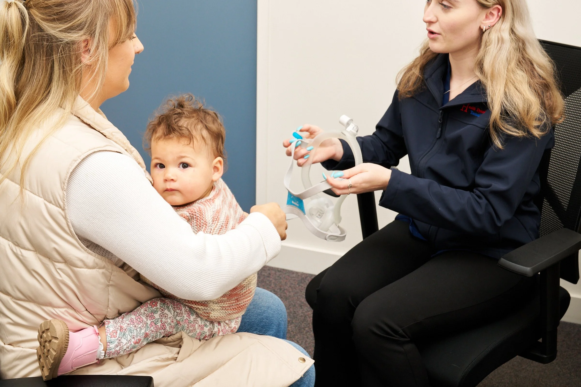 A young child in a pink sweater and patterned leggings sitting on a woman's lap in a medical office, with a healthcare professional holding a mask.