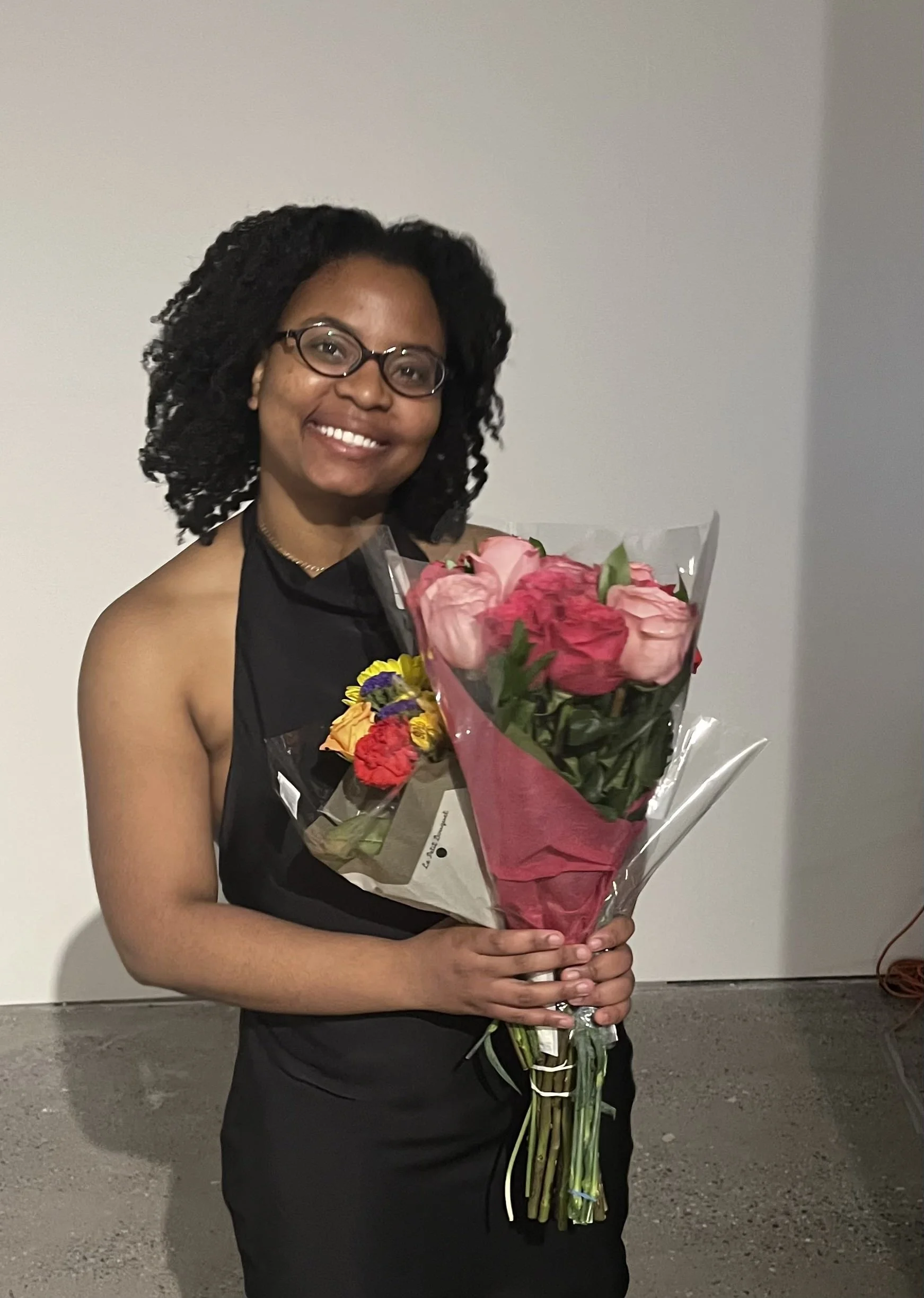 A woman with short curly hair, glasses, and a black dress is smiling and holding a bouquet of pink and yellow flowers. She is standing against a plain gray wall.