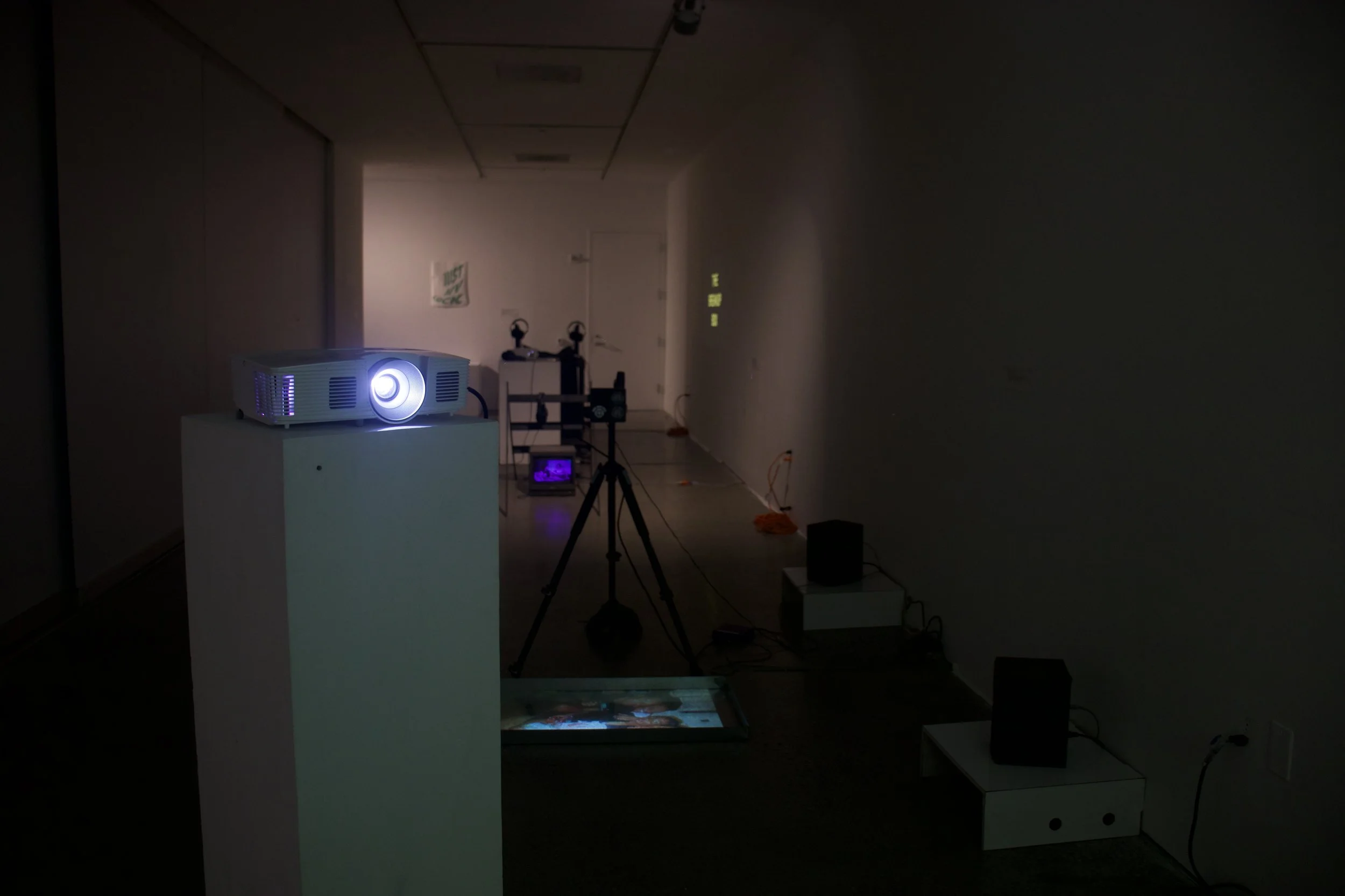 A dimly lit room with a projector on a white pedestal casting a light on the wall. There's a tripod and various electronic equipment, including a small monitor, on the floor and tables. The walls are mostly plain with some small items and signs, and 