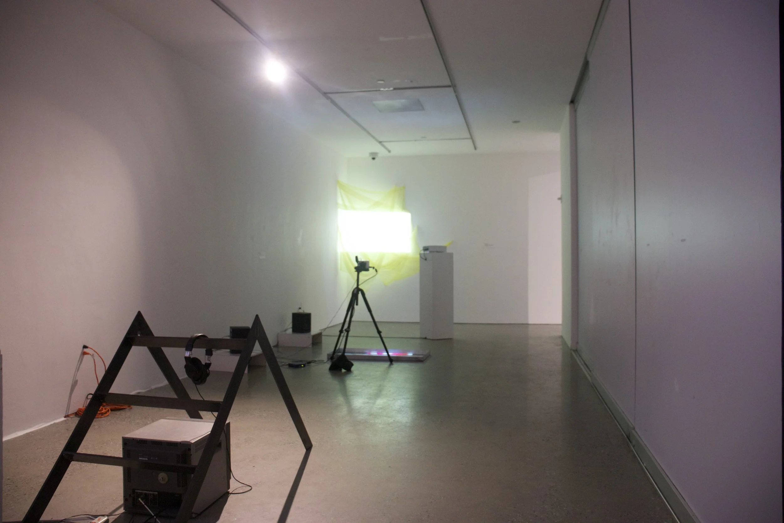 An art gallery or exhibition space with white walls and a polished floor, featuring a few technical apparatuses such as a tripod with a camera and a projector, and a yellow fabric installation covering a window with bright light shining through.