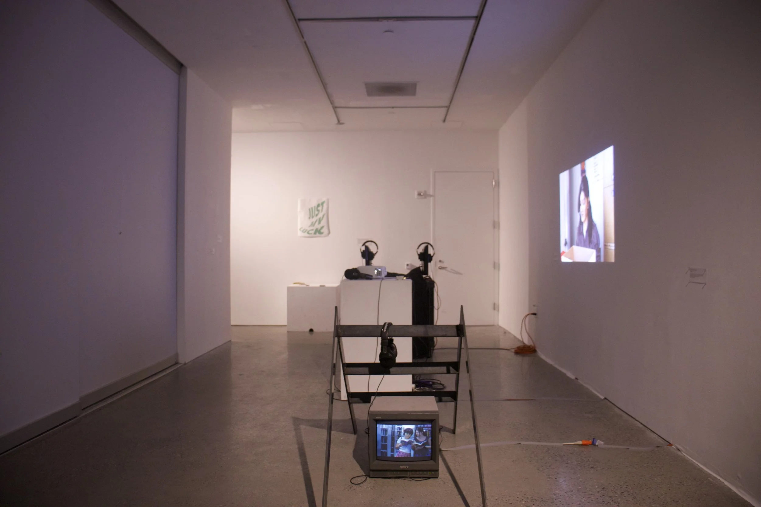 An art installation featuring a TV screen on a stand displaying two children reading books, with a smaller TV below showing a similar image, in a gallery space with white walls and minimal decor.