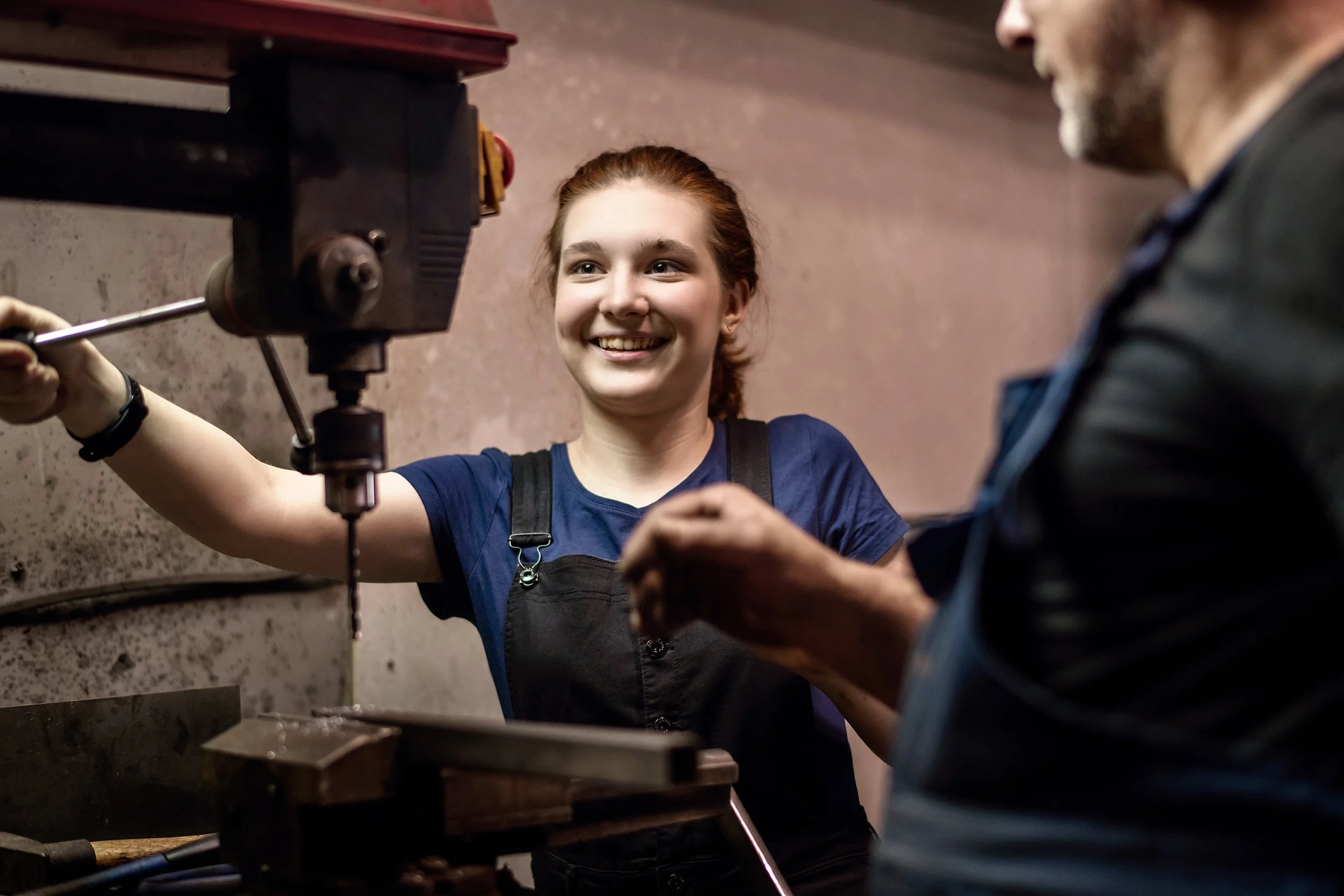 A young woman with red hair smiling while operating a drill press in a workshop, assisted by an older man.