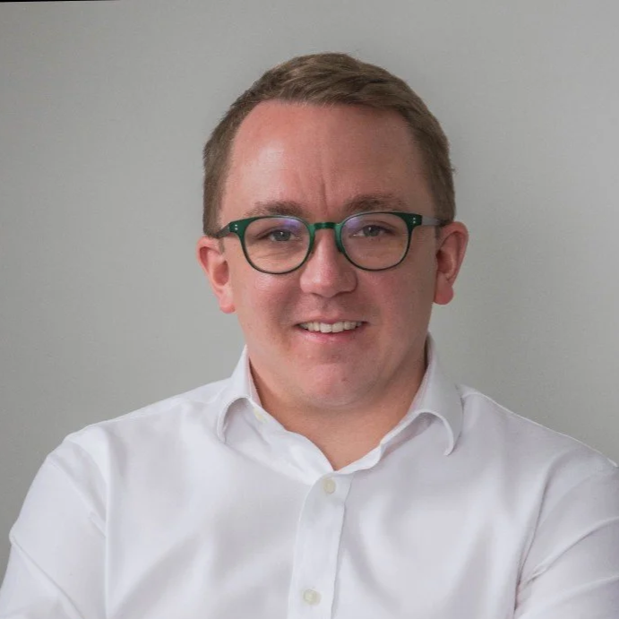 A young man with short brown hair, wearing glasses with green frames and a white button-up shirt, smiling at the camera against a plain gray background.