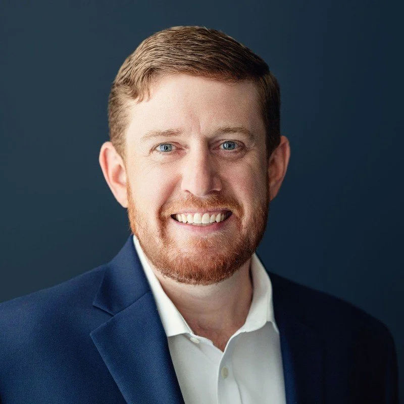 Headshot of a man with red hair and beard, wearing a navy suit and white shirt, smiling at the camera against a dark background.