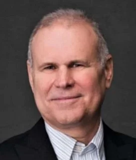 Portrait of a middle-aged man with short, light-colored hair, wearing a dark suit and tie, against a dark background.