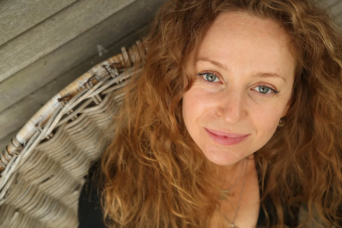 Ilana Laps, a woman with curly red hair and blue eyes smiling, lying on a woven outdoor chair with a wooden wall in the background.
