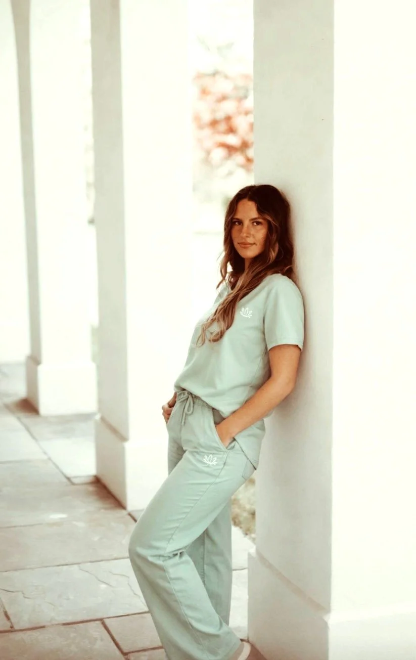 A man with dark, curly hair, glasses, and facial hair, looking to the side, wearing a dark sweater, with a plant and light-colored wall in the background.