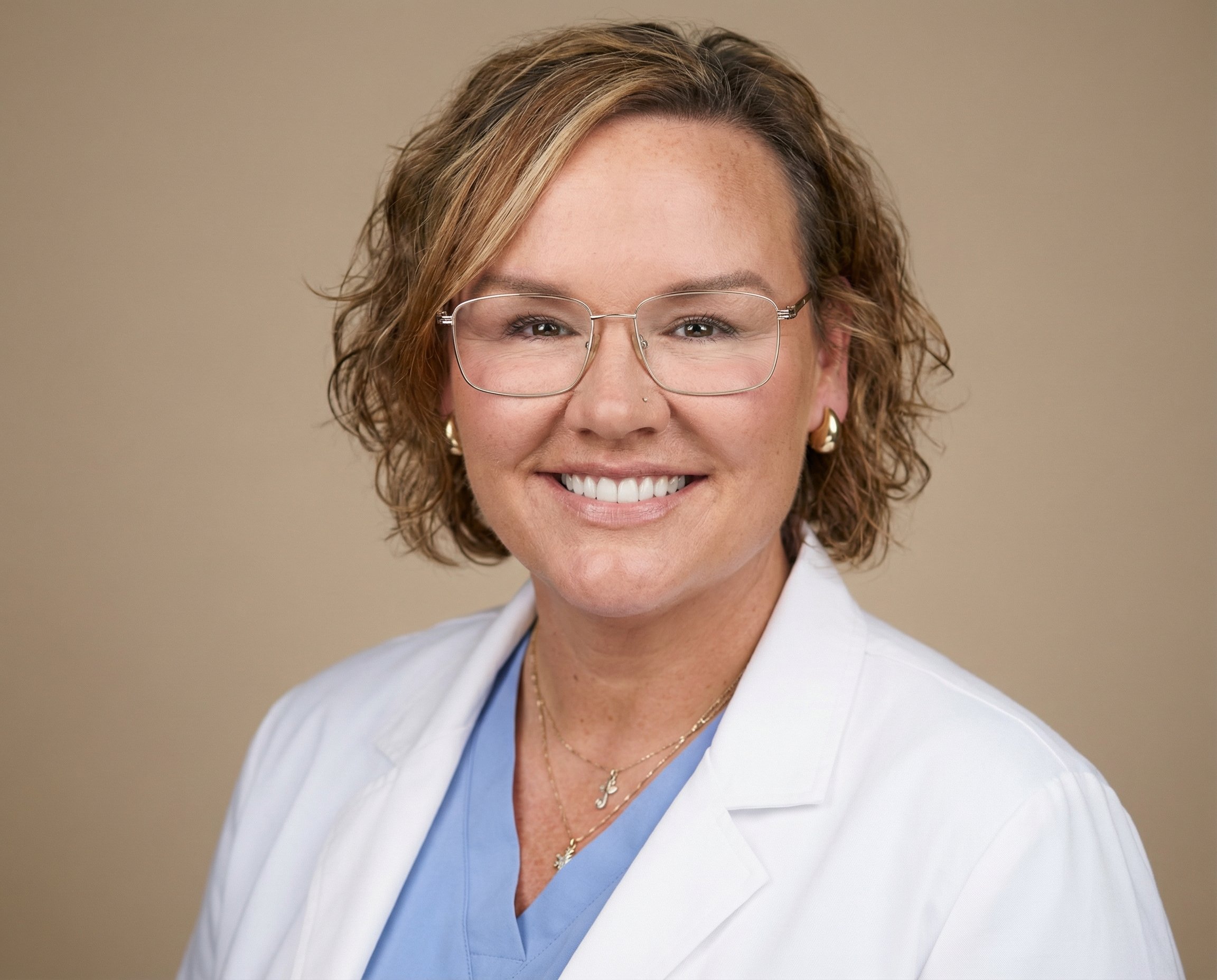 A smiling woman with curly black hair, wearing glasses, earrings, and a striped shirt, indoors with a blurred background.