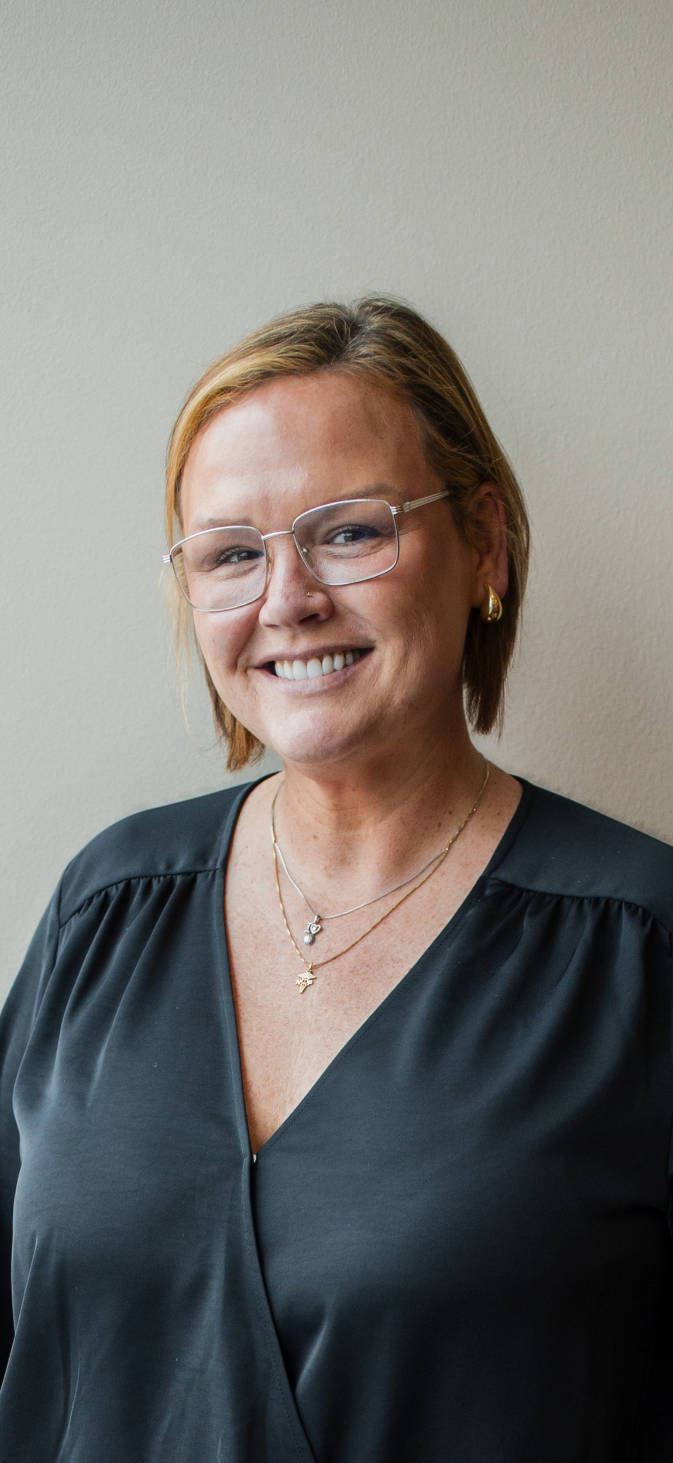 A smiling woman with curly black hair, wearing glasses, earrings, and a striped shirt, indoors with a blurred background.