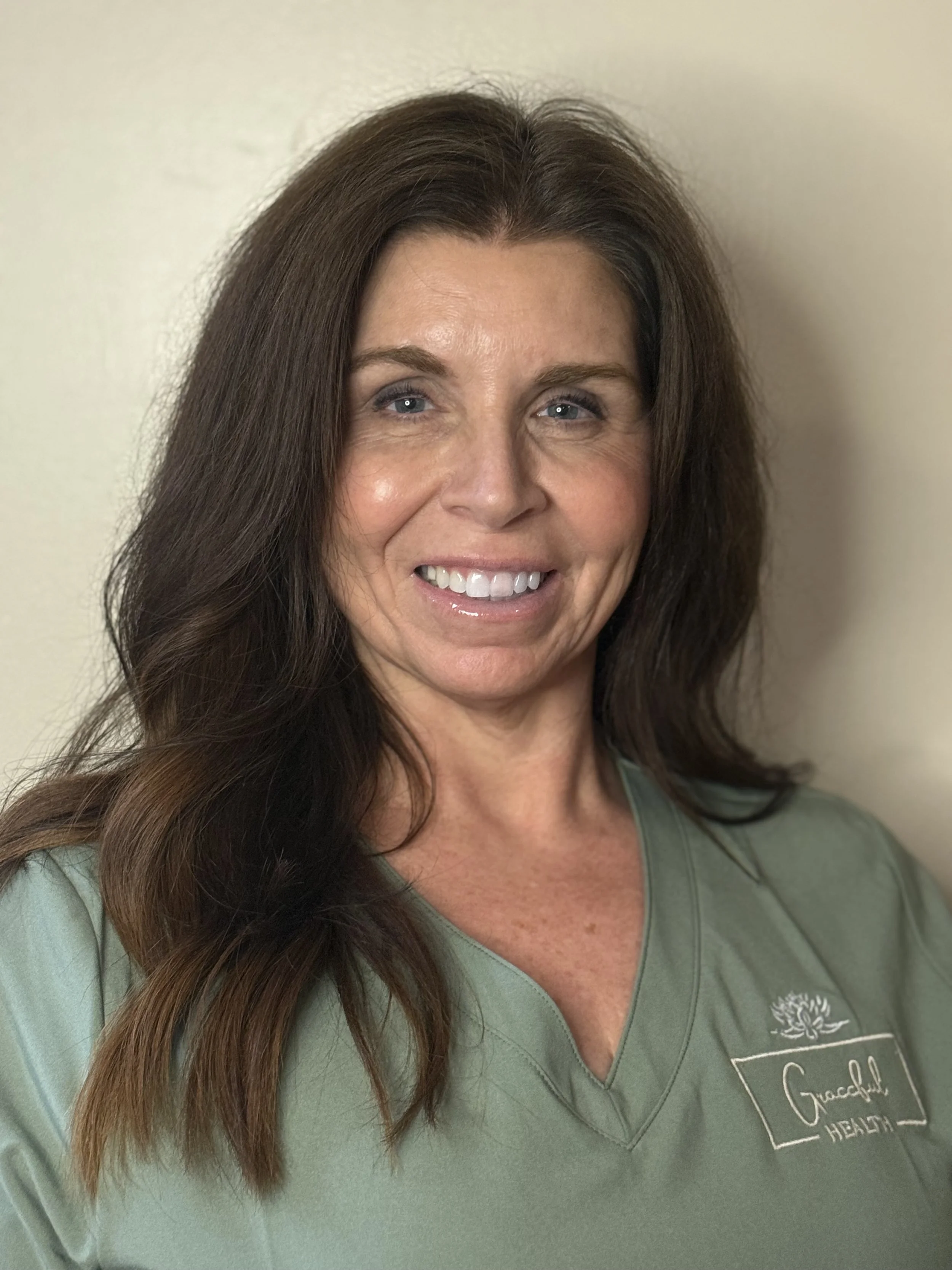 Smiling woman with shoulder-length black hair, wearing a blue top, surrounded by green plants.