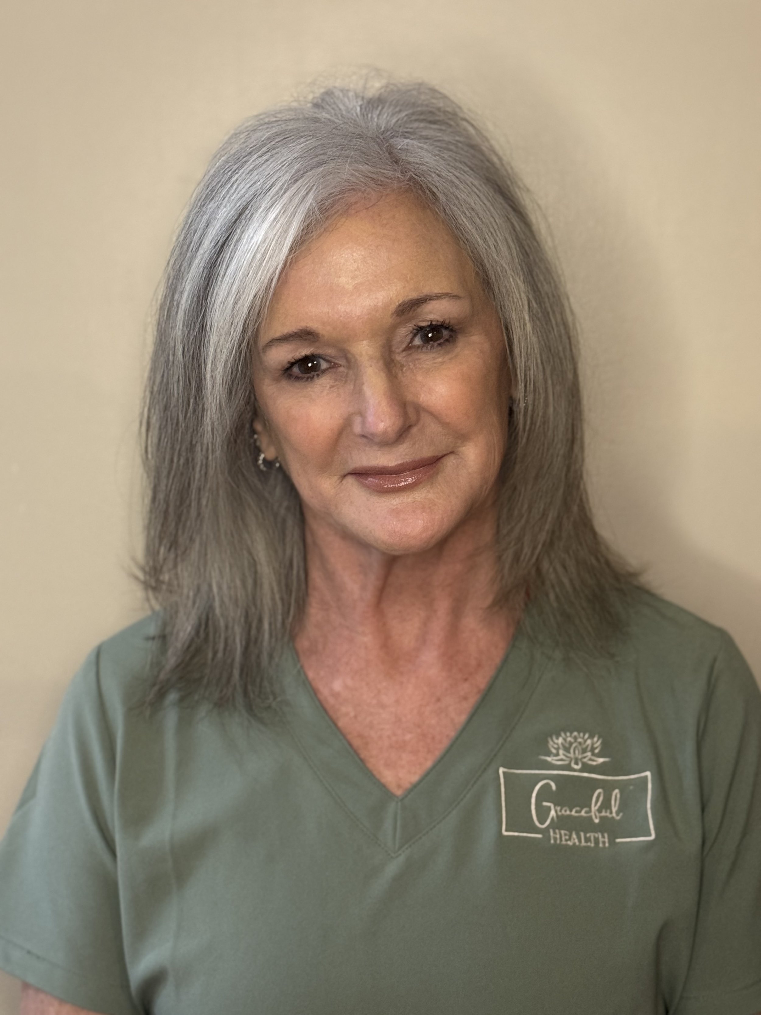 A smiling woman with gray hair wearing a green medical uniform with the logo 'Graceful Health'.