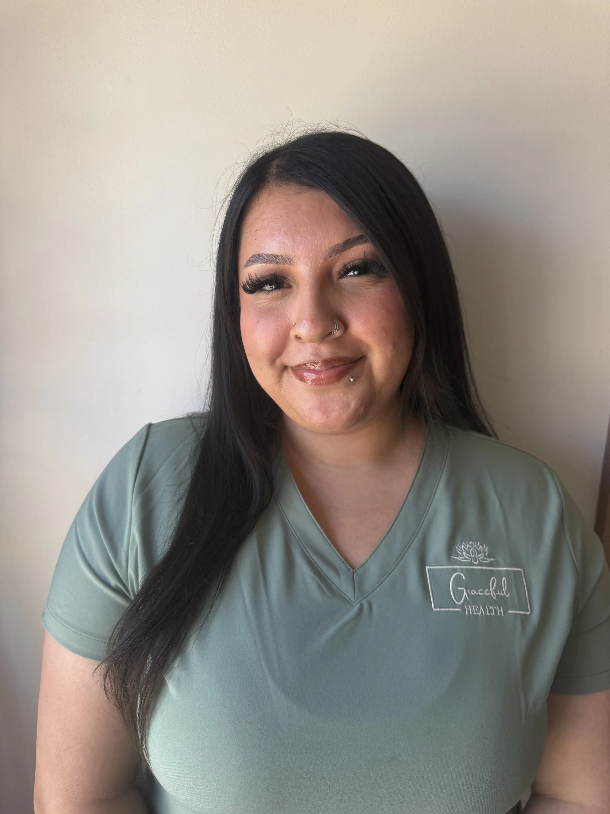 A woman with long black hair, wearing makeup, a nose and lip piercing, and a green health care uniform with a logo and name tag that says 'Graceful Health' are smiling in front of a plain light-colored wall.