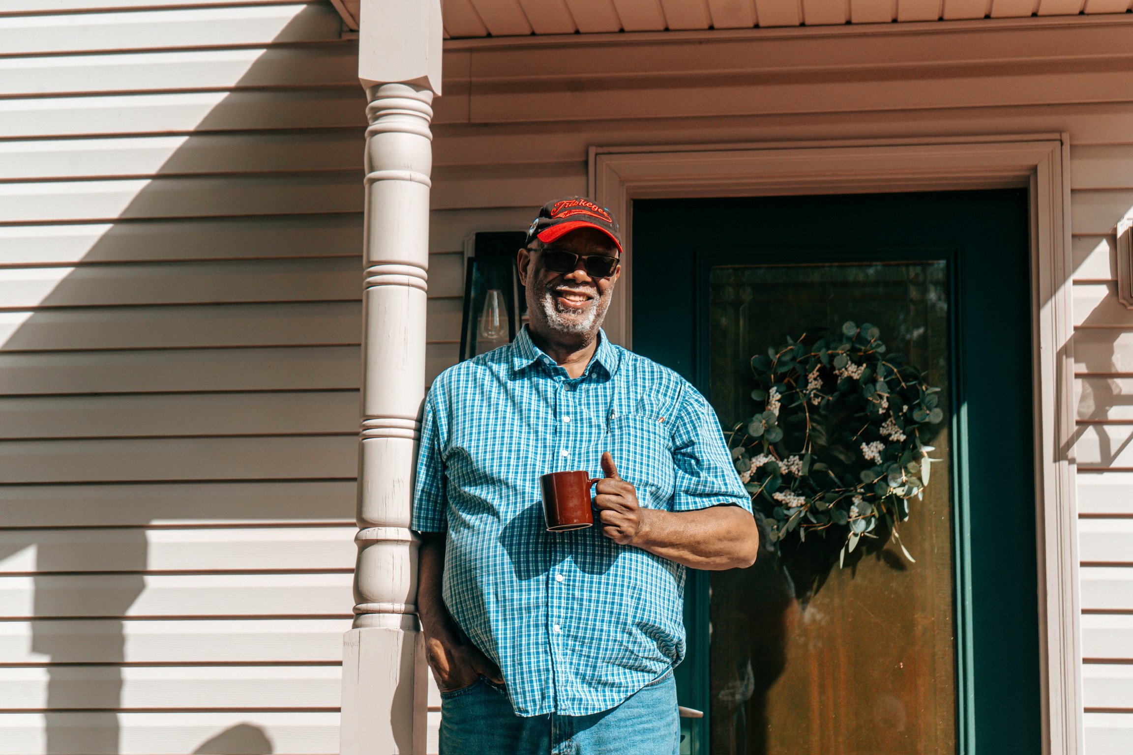 A cheerful older man with gray hair and a beard stands outside a house, smiling and giving a thumbs up. He is wearing sunglasses, a red cap, a blue plaid shirt, and holding a red mug.