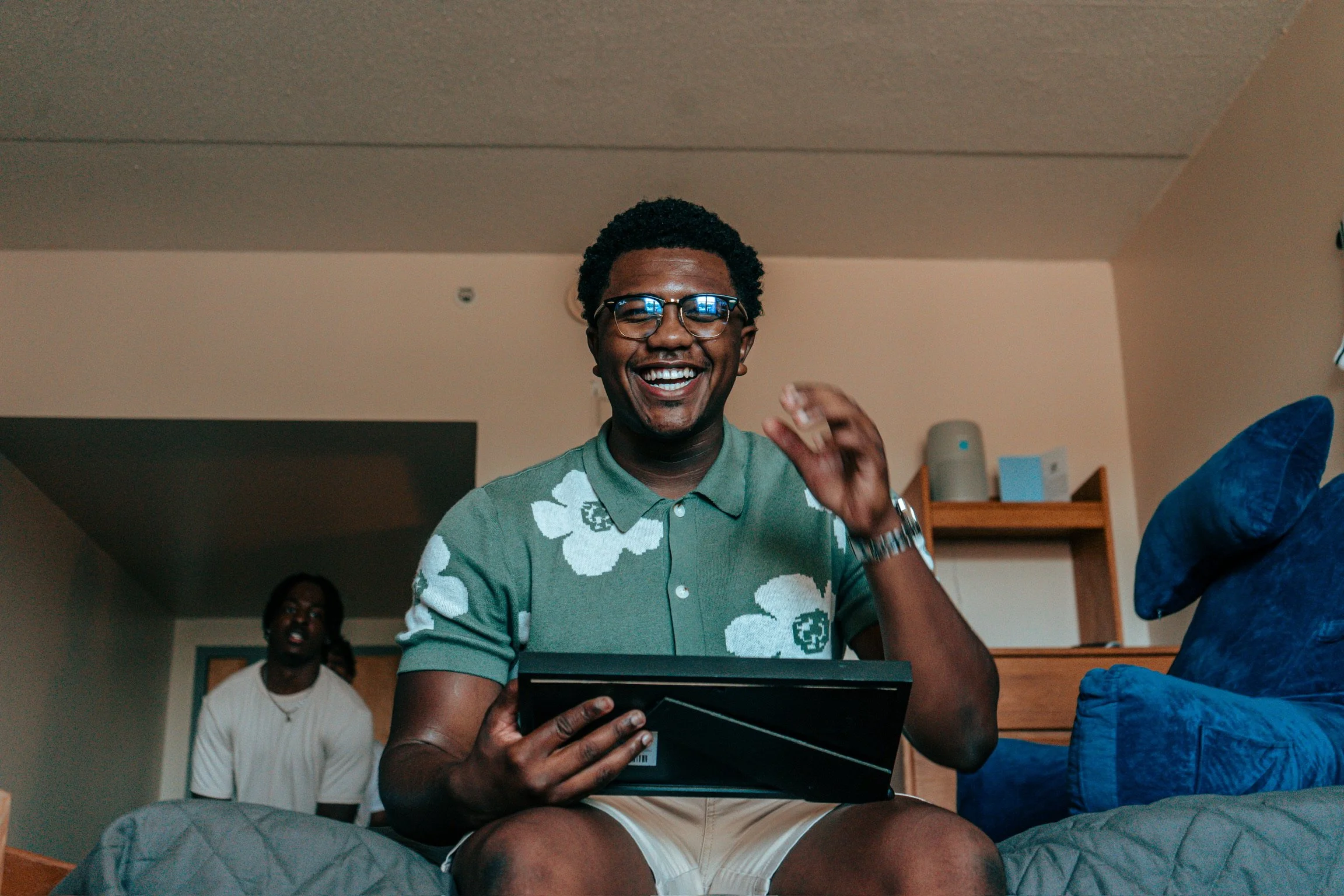 A man with glasses smiling and holding a tablet, sitting on a bed in a cozy apartment, with a woman in the background.