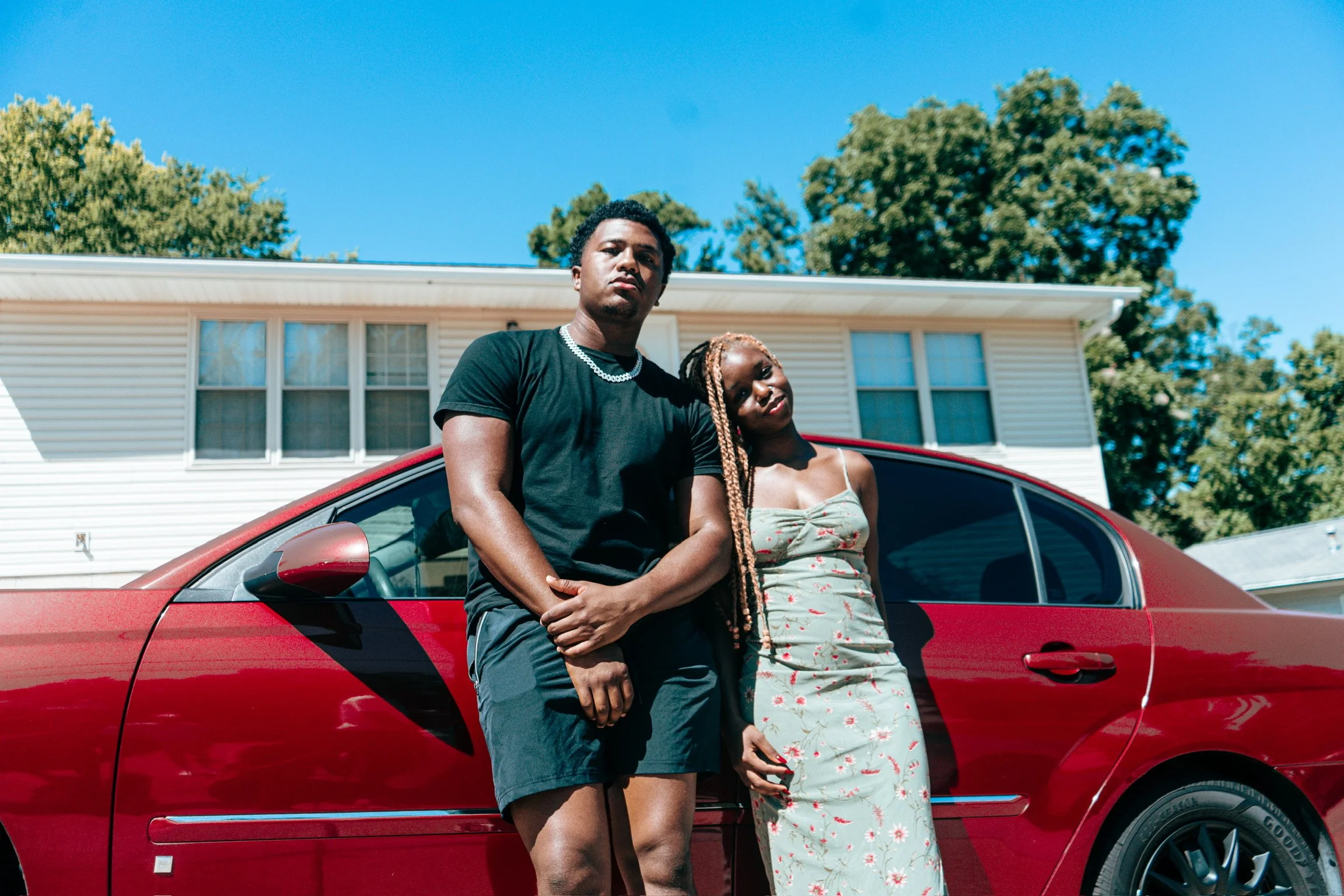 A young Black man and woman are leaning against a red car parked in front of a white house with a blue sky background.